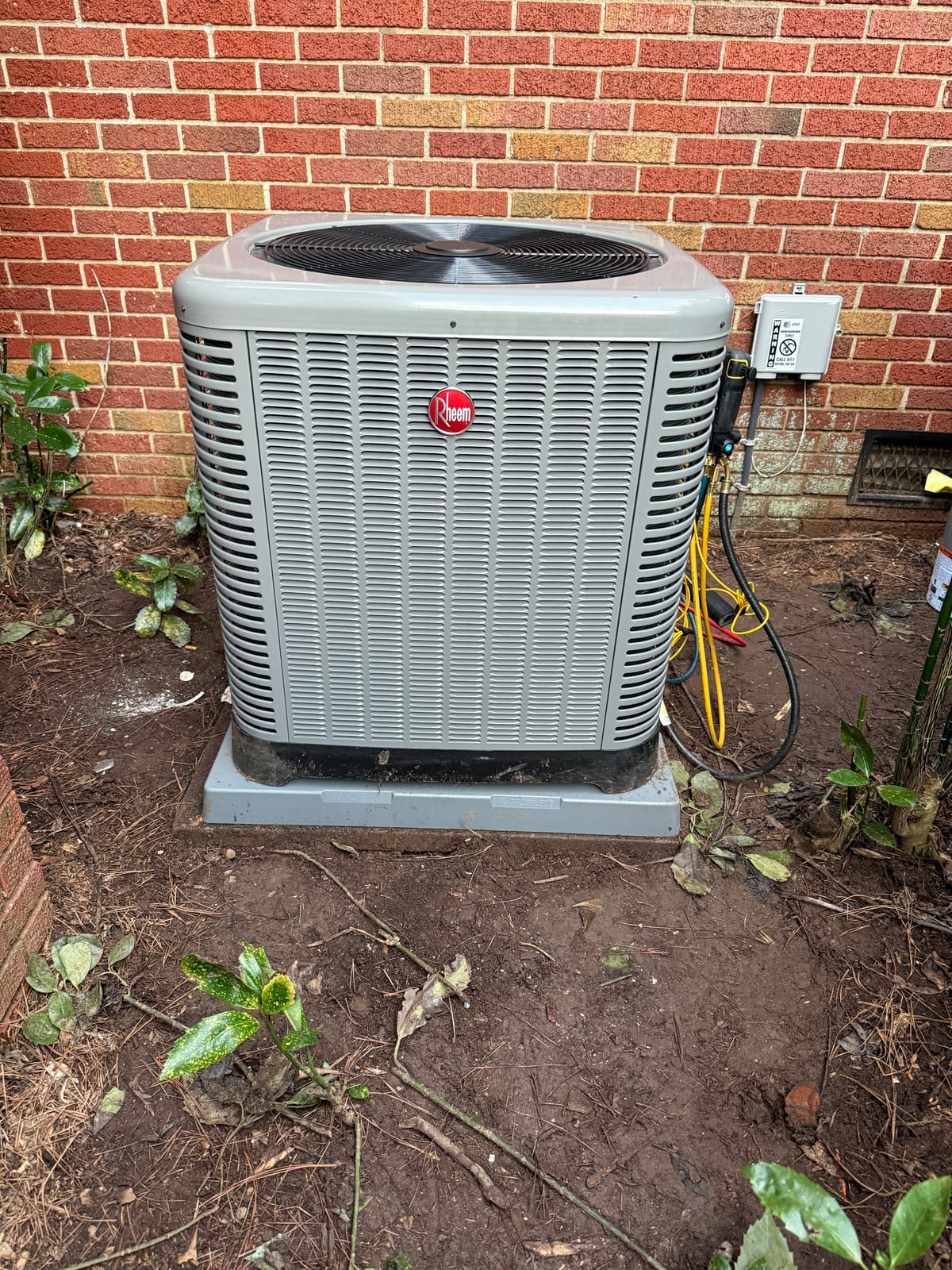First Class technician commissioning a new Rheem AC condenser on a pad against a brick home with refrigerant gauges connected